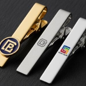 A variety of elegant custom logo tie clips in gold and silver finishes, arranged on a dark, textured surface.