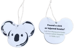 Two koala-head shaped custom air fresheners. The front view shows a black and white graphic of a koala's face. The back view displays black text on a white background: 'Found a sick or injured koala? Call Port Macquarie Koala Conservation Hospital 02 6584 1522'. Both have white hanging strings.