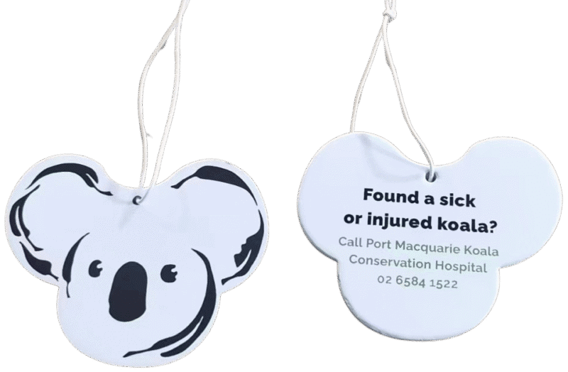 Two koala-head shaped custom air fresheners. The front view shows a black and white graphic of a koala's face. The back view displays black text on a white background: 'Found a sick or injured koala? Call Port Macquarie Koala Conservation Hospital 02 6584 1522'. Both have white hanging strings.