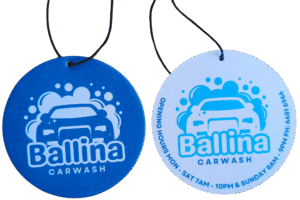 Two round custom air fresheners for 'Ballina CARWASH'. One version is dark blue with a white logo (car silhouette amid bubbles) and text. The other is white with the same logo and 'Ballina CARWASH' text in blue, also including 'OPENING HOURS MON - SAT 7AM - 10PM & SUNDAY 8AM - 9PM PH: 6681 6966' in blue around the perimeter. Both have black hanging strings.