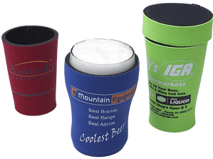 Three neoprene stubby holders in red, blue, and green, designed as branded promotional items, are displayed against a light grey background. The red holder on the left features a partially visible gold text logo. The blue holder in the center, shown with a white can inside, displays a "Mountain Equipment" logo with the text "Best Brands Best Range Best Advice" and "Coolest Beer!". The green holder on the right, with a black base and trim, is printed with "Kelly's IGA Supermarkets" and contact details, along with a liquor advertisement. These cylindrical sleeves are intended to insulate beverage cans or bottles.